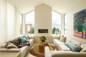 Coastal living room with central brick fireplace, wraparound built-in seating, soft neutral palette and ocean views in the Cottage by the Sea Charity Home, designed for relaxed, communal living.