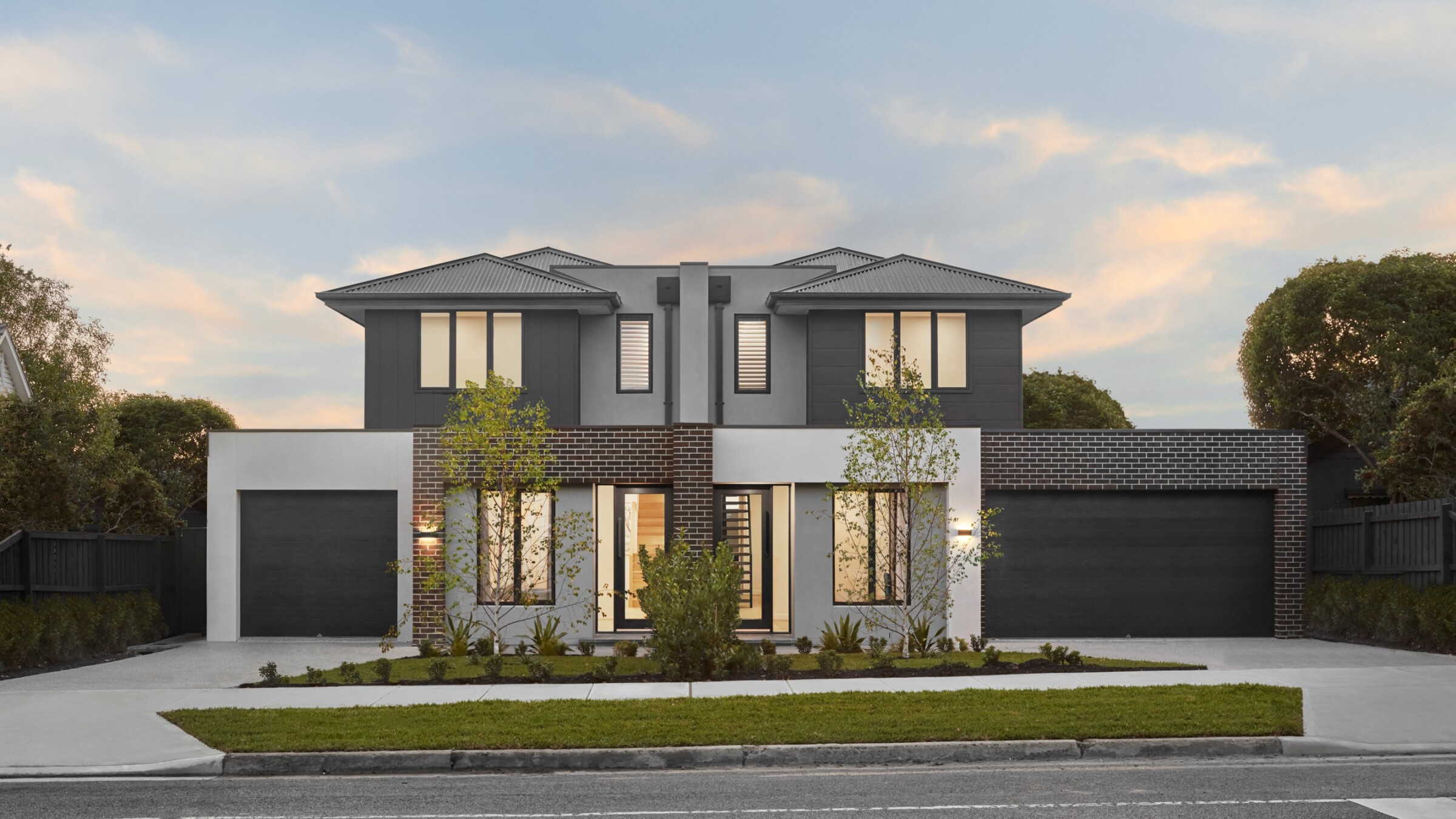 Modern two-story duplex house with symmetrical design, featuring dark gray and white exterior, brick accents, large windows, and double garages on a quiet suburban street at sunset.